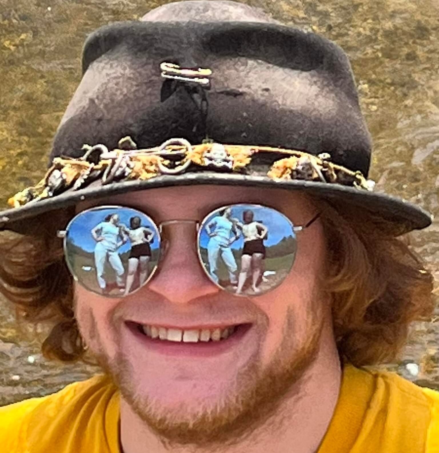 Talon Adastra the man behind the Mischief Juggling movement. In a black cap and a yellow shirt, smiling and wearing reflective sunglasses with circus actors in the background.